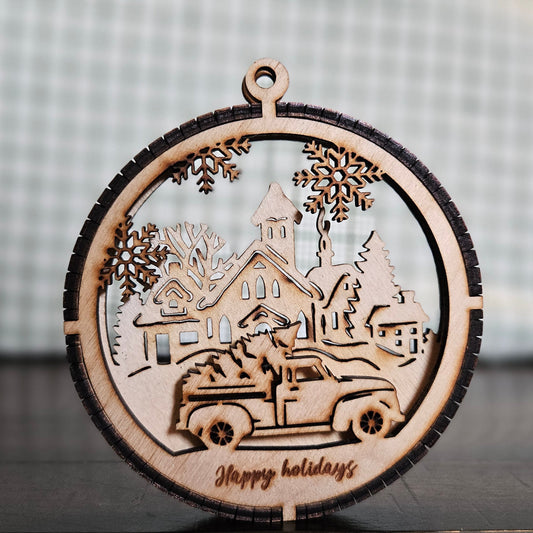 A two-layer wooden ornament featuring a truck carrying a Christmas Tree in the foreground and a winter village scene in the background.