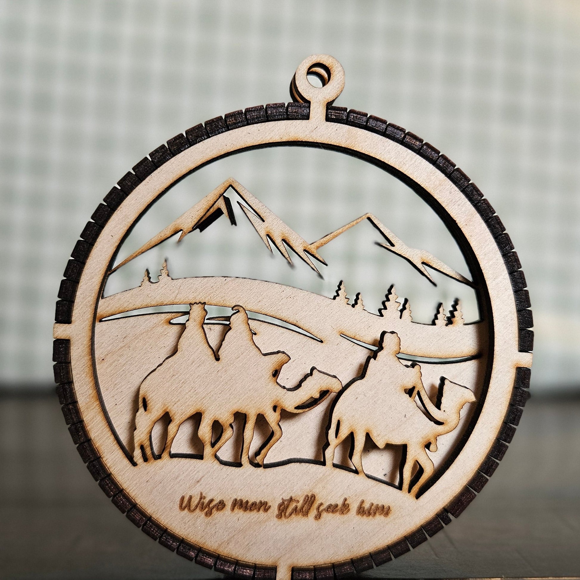 A two-layer wooden ornament featuring the 3 wise men on camel-back in the foreground and a mountain scene in the background.